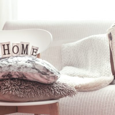 Home decoration. Decorative pillows and home lettering on white wooden stylish chair. Sofa and wicker basket with cushions in the background