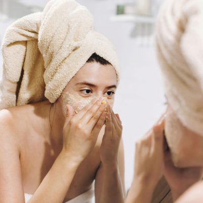Young happy woman in towel making facial massage with  organic face scrub and looking at mirror in stylish bathroom. Girl applying scrub cream, peeling and cleaning skin. Skin Care