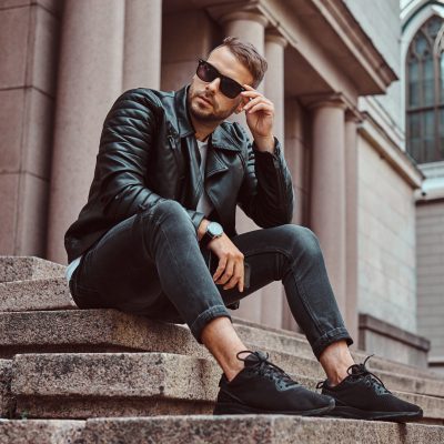 Fashionable guy dressed in a black jacket and jeans holds a smartphone sitting on steps against an old building in Europe.