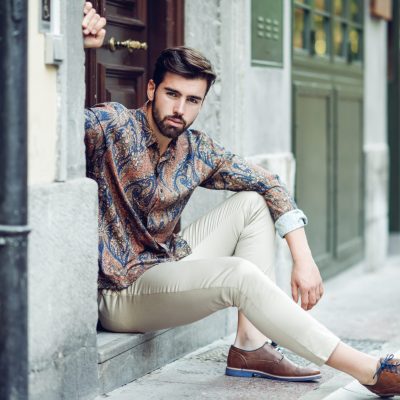 Young bearded man, model of fashion, sitting in an urban step wearing casual clothes. Guy with beard and modern hairstyle looking away in the street.